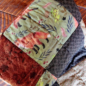 May include: A quilted fabric item featuring a patchwork design with panels of sage green fabric printed with red foxes, leaves, and berries, alternating with dark brown fabric with small floral patterns and a rust-colored section with leaf motifs.