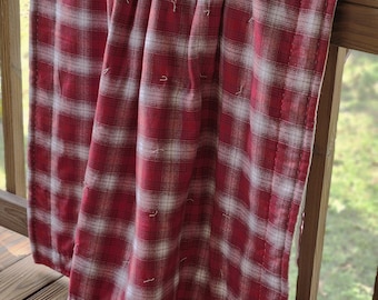 May include: A rectangular quilt featuring a red and white plaid pattern with visible white hand-stitched quilting details throughout the fabric. The textile is draped over a wooden railing, showcasing its soft texture and classic checkered design.