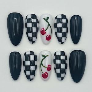 May include: A set of ten oval-shaped false nails. The nails feature a variety of designs, including solid black, black and white checkered patterns, and white nails with red cherry designs.