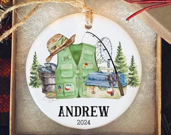 May include: A round, white ceramic Christmas ornament with a green fishing vest, a fishing rod, a bucket, and a fishing lure. The ornament is personalized with the name "ANDREW" and the year "2024".