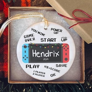 May include: A white round Christmas ornament with a blue and red Nintendo Switch controller design. The ornament has the text "YOU WIN!" "GAME OVER" "POWER UP" "START UP" "Hendrix 2024" "PLAY 1 PLAYER 2 PLAYER" "HI-SCORE" "SAVE" "OK" printed on it.