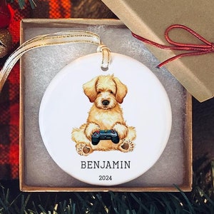 Puede incluir: Adorno navideño de cerámica blanca con una ilustración de dibujos animados de un caniche toy sosteniendo un mando de videojuegos. El adorno está personalizado con el nombre "Benjamin" y el año "2024".