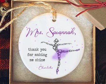 Puede incluir: Adorno de cerámica blanca con un diseño de bailarina y el texto "Mrs. Savannah, thank you for making me shine - Charlotte." El adorno está en una caja de regalo con una cinta roja, rodeado de ramas de árbol de Navidad.