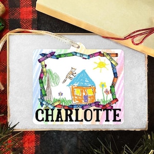 Może przedstawiać: Biała ceramiczna ozdoba choinkowa z kolorową ramką z kredek. Wewnątrz ramki znajduje się rysunek dziecka przedstawiający dom, kota, drzewo i słońce. Ozdoba jest spersonalizowana imieniem "CHARLOTTE".