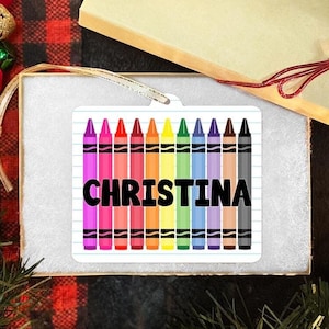 Może przedstawiać: Biała ozdoba z obrazkiem kredek w kolorach tęczy i imieniem Christina napisanym czarnymi literami na kartce w linie.