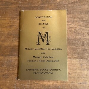 May include: A gold-coloured booklet titled "CONSTITUTION and BYLAWS of Midway Volunteer Fire Company and Fireman's Relief Association." The booklet is approximately 10 cm long, with black text. The text includes the location: Lahaska, Bucks County, Pennsylvania.