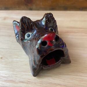 Könnte beinhalten: Ein brauner Aschenbecher aus Keramik in Form eines Hundekopfes mit roten und weißen Details. Der Hund hat den Mund offen und das Innere ist hohl.