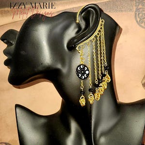 May include: Gold ear cuff with a black and white moon phase charm and dangling chains with black beads. The ear cuff is on a black mannequin head.