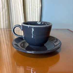 May include: A dark gray ceramic teacup and saucer set. The cup has a curved handle and sits on a matching saucer. The set is placed on a reflective, brown surface.