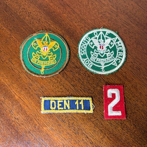 May include: Four vintage Boy Scout patches. Two circular patches with the Boy Scouts of America logo and the words "Be Prepared". A rectangular patch with "DEN 11" and a red patch with the number "2".