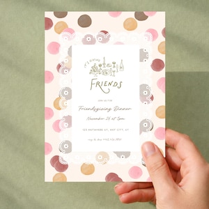 Op de afbeelding: Een rechthoekige uitnodigingskaart met een crèmekleurige achtergrond en verspreide roze, bruine en beige stippen. Een wit centraal paneel met een kanten rand toont de tekst: It's Giving Friends, Join us for Friendsgiving Dinner, November 26 at 5pm, 123 Anywhere St, Any City, ST, rsvp to Jane 123.456.7890.