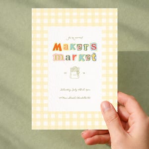 May include: A rectangular invitation card featuring a light yellow and white gingham border. The center white area displays the text "You're invited! Makers market" in colorful, playful letters, a line drawing of a tote bag, and event details.