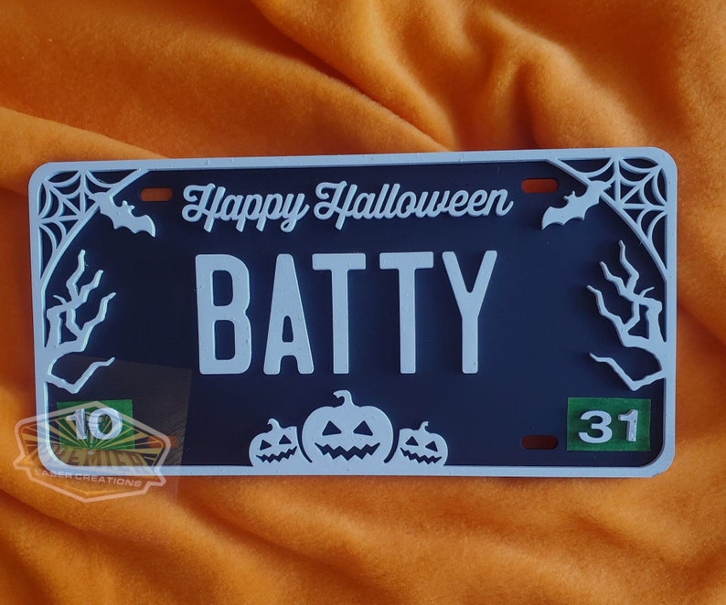 May include: A black and white Halloween-themed license plate with the text "Happy Halloween BATTY" and a spiderweb design. The plate also features three white pumpkins with jack-o'-lantern faces and the numbers "10" and "31" in green.