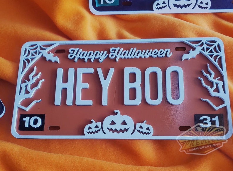 May include: A brown and white Halloween license plate with the text "Happy Halloween" and "HEY BOO". The plate is decorated with a spider web, bats, and pumpkins.