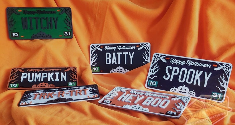 May include: Five Halloween-themed license plate style signs with black, white, orange, and green paint. The signs feature the words "Witchy", "Pumpkin", "Batty", "Spooky", and "Hey Boo" with a spiderweb and bat design.