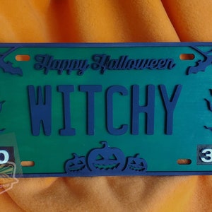 May include: Green and blue Halloween-themed license plate with the text "Happy Halloween" and "Witchy" in a dark blue font. The plate has a spiderweb design and two bats on either side of the text. The numbers "10" and "31" are on the left and right sides of the plate, respectively. There are three carved pumpkins at the bottom of the plate.