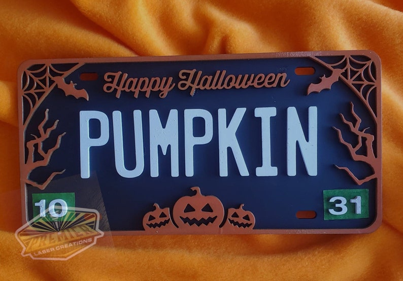 May include: A blue and copper Halloween license plate with the text "Happy Halloween PUMPKIN" and three orange pumpkins with jack-o'-lantern faces. The numbers 10 and 31 are in the corners of the plate.