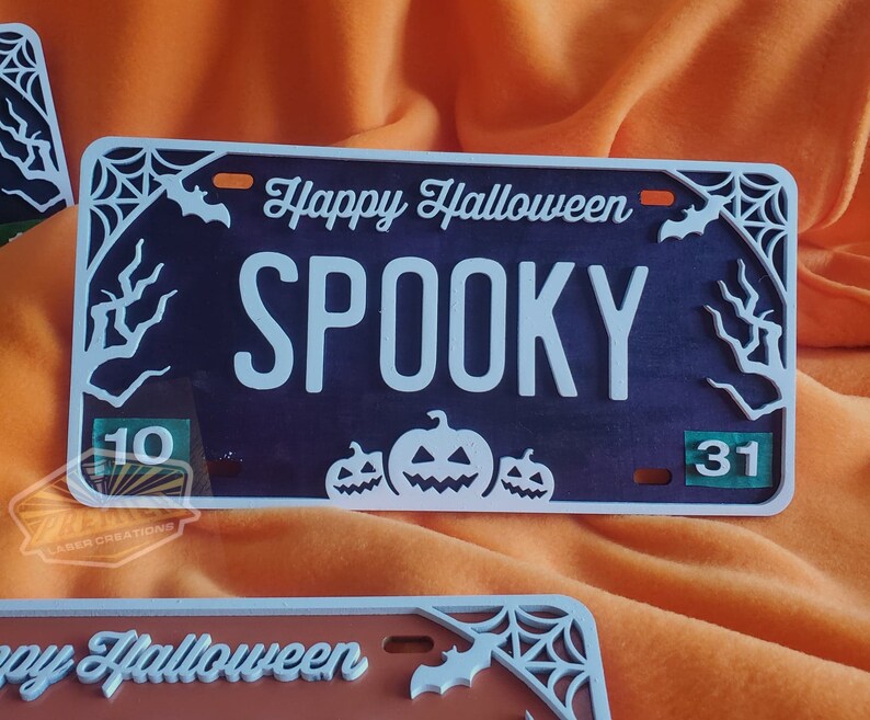 May include: A black and white Halloween license plate with the text "Happy Halloween" and "SPOOKY" in white. The plate has a spiderweb design with bats and a green number "31" in the corner. There are three white pumpkins with orange faces in the bottom center.