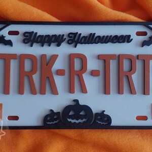 May include: A white Halloween-themed license plate with black trim and orange lettering. The plate reads "Happy Halloween" and "TRK-R-TRT". The numbers "10" and "31" are on either side of the plate. There are three black pumpkin silhouettes at the bottom of the plate.