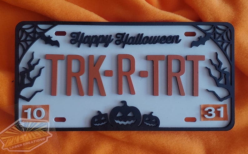 May include: A white Halloween-themed license plate with black trim and orange lettering. The plate reads "Happy Halloween" and "TRK-R-TRT". The numbers "10" and "31" are on either side of the plate. There are three black pumpkin silhouettes at the bottom of the plate.