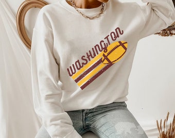 Può includere: Felpa bianca con un design a strisce marroni e dorate e una grafica di un pallone da calcio. Il testo "WASHINGTON" è stampato sulla felpa.