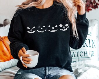 May include: Black sweatshirt with a row of five white pumpkin faces. The sweatshirt is worn by a person with long blonde hair and is sitting on a white couch.