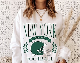 May include: White sweatshirt with a green and white graphic design that says "New York Football Est. 1960" with a football helmet graphic.