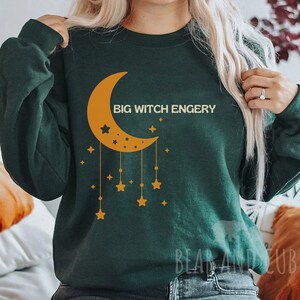 以下が含まれることがあります： 三日月と星のグラフィックが描かれた濃い緑色のスウェットシャツ。スウェットシャツには「BIG WITCH ENGERY」という文字がプリントされています。