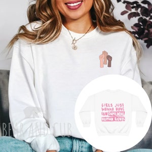 May include: White sweatshirt with the text "Girls just wanna have fundamental human rights" in pink lettering. The sweatshirt also features a graphic of three raised fists in different skin tones.