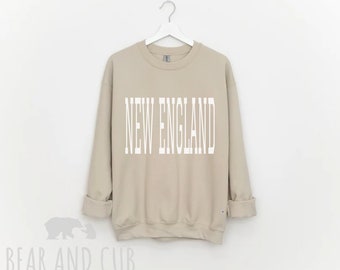 Può includere: Una felpa beige con la scritta "NEW ENGLAND" stampata in bianco sul davanti.