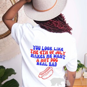 May include: White t-shirt with red, white, and blue text that reads "You look like the 4th of July makes me want a hot dog real bad". A red and white hot dog is pictured below the text.