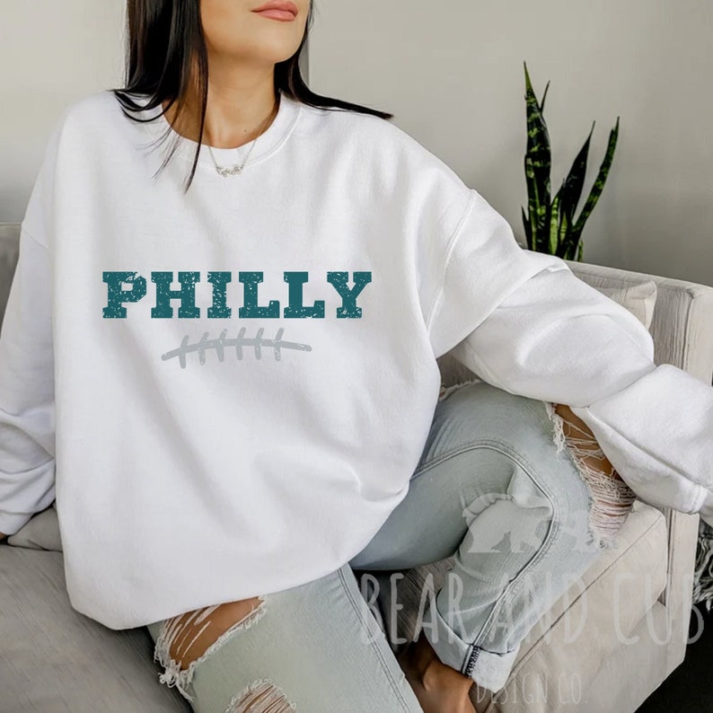 Pu&ograve; includere: Felpa bianca con la parola "PHILLY" in lettere verde acqua dall'aspetto usurato. La parola &egrave; sopra un disegno di pallone da calcio cucito grigio.
