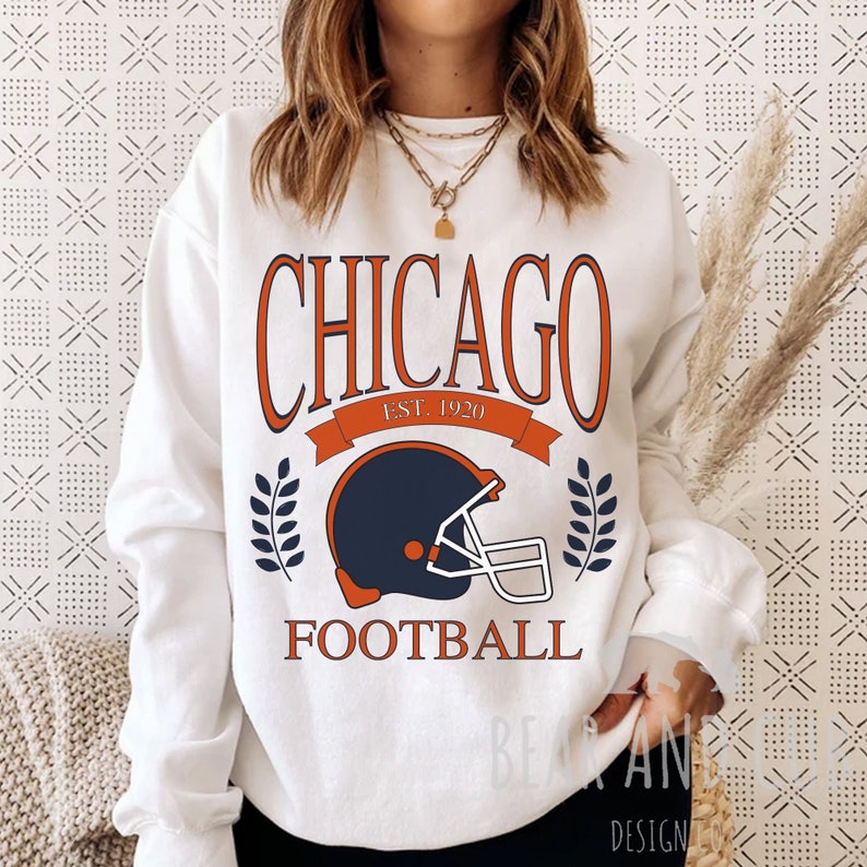 May include: White sweatshirt with a graphic design featuring the text "CHICAGO EST. 1920 FOOTBALL" in orange and blue, with a blue and white football helmet graphic.