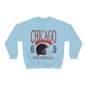 May include: Light blue sweatshirt with a vintage-style graphic of a football helmet and the text "CHICAGO EST. 1920 FOOTBALL".