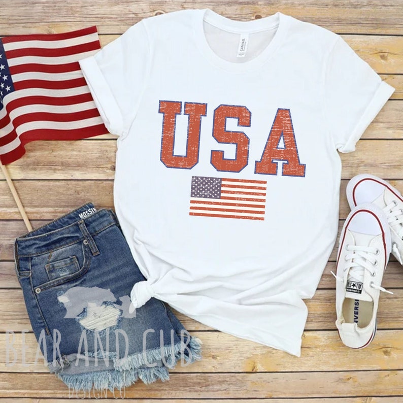 May include: White t-shirt with a distressed red and blue "USA" graphic and a small American flag.