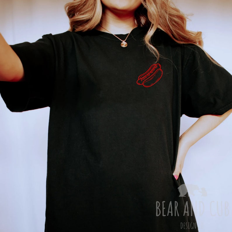 May include: A black t-shirt with a red outline of hot dogs on the front. The shirt is being worn by a person.