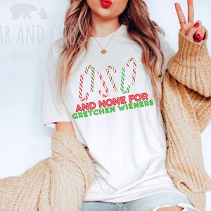 以下が含まれることがあります： キャンディーケーンのグラフィックと「AND NONE FOR GRETCHEN WIENERS」というテキストが書かれた白いTシャツ。