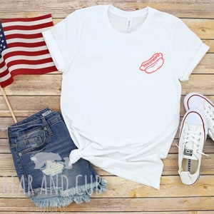 May include: White t-shirt with a red outline of a hot dog on the left chest. The shirt is laying on a wooden surface with a pair of blue denim shorts, a small American flag, and a pair of white sneakers.