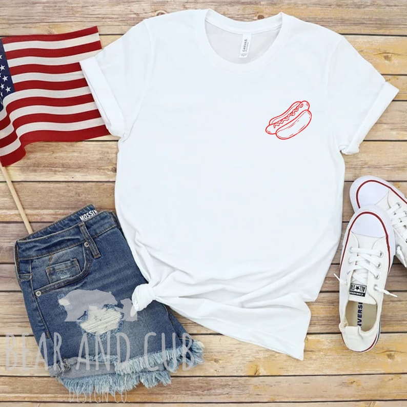 May include: White t-shirt with a red outline of a hot dog on the left chest. The shirt is laying on a wooden surface with a pair of blue denim shorts, a small American flag, and a pair of white sneakers.