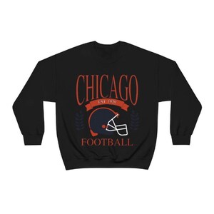 May include: Black sweatshirt with a red and white graphic design that says "CHICAGO EST. 1920 FOOTBALL" with a blue and orange football helmet in the center.