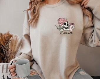 Può includere: Una felpa beige chiaro con una grafica di uno scheletro che indossa un berretto rosa e tiene una tazza di caffè. Il testo "STAYIN' ALIVE" è stampato sotto la grafica.