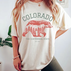 Puede incluir: Una camiseta de color crema con un gr&aacute;fico de silueta de oso rojo. El oso camina por un bosque con monta&ntilde;as y estrellas en el fondo. El texto "COLORADO" est&aacute; en verde sobre el oso y "THE CENTENNIAL STATE EST. 1876" est&aacute; debajo del oso.