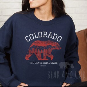 May include: Navy blue sweatshirt featuring a red graphic of a bear with mountains and trees inside its silhouette. The text "COLORADO" is above the bear and "THE CENTENNIAL STATE EST. 1876" is below the bear.