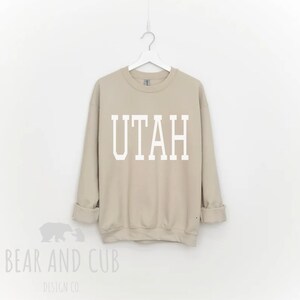 May include: A tan sweatshirt with the word "UTAH" printed in white block letters. The sweatshirt is hanging on a white hanger against a white background. The Bear and Cub Design Co. logo is visible in the bottom left corner.