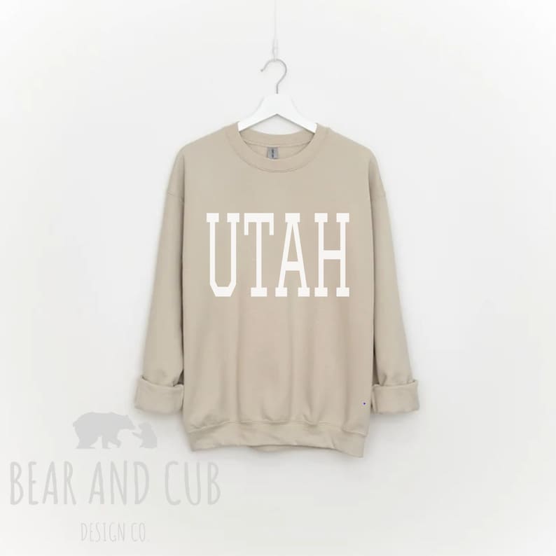 May include: A tan sweatshirt with the word "UTAH" printed in white block letters. The sweatshirt is hanging on a white hanger against a white background. The Bear and Cub Design Co. logo is visible in the bottom left corner.