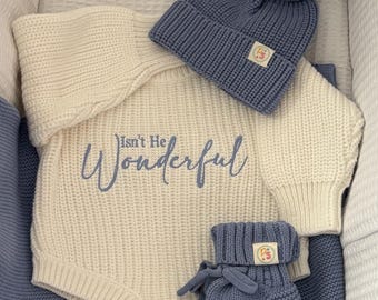 May include: A baby outfit set in a white bassinet. The set includes a cream-colored romper with "Isn't He Wonderful" embroidered in blue, a blue knit hat with a pom-pom, and matching booties. A blue blanket is also included.
