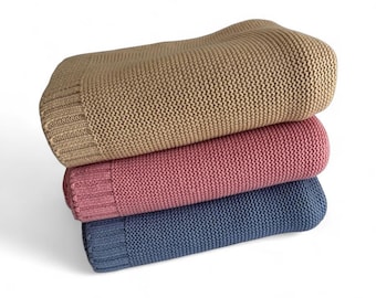 May include: Three folded knit blankets in a stack. The top blanket is tan, the middle is pink, and the bottom is blue. Each blanket has a ribbed border at the end. The blankets are made of a thick, textured knit.