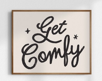 May include: A framed print with a white background and black text that reads "Get Comfy" with two small stars on either side of the text.