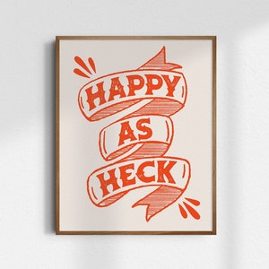 May include: A framed print with a white background and red text that reads "Happy as Heck" in a stylized font. The text is on a red ribbon that is curled and tied in a knot.