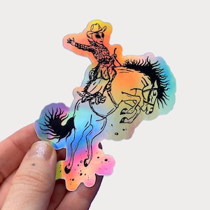 Peut inclure: Un autocollant holographique repr&eacute;sentant une illustration en noir et blanc d'un extraterrestre portant un chapeau de cowboy chevauchant un cheval cabr&eacute;.